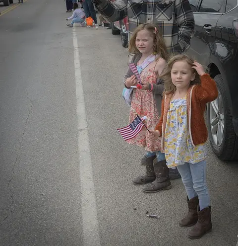 Two young girls holding American flags stand by the side of a road during an event.