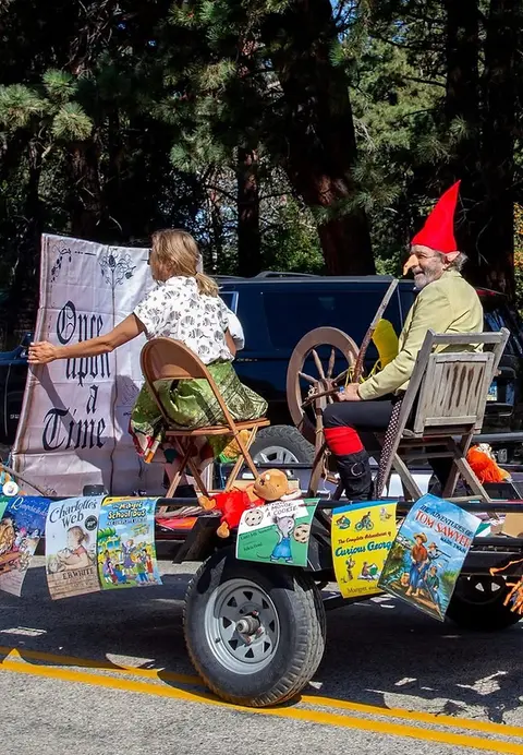 Two people dressed in costume ride on a parade float decorated with book covers.