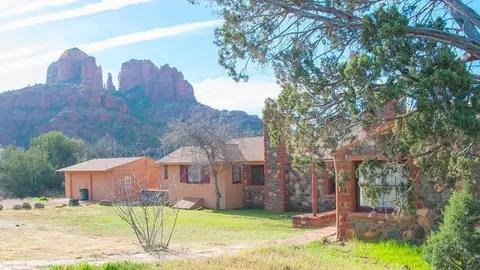 A rustic stone and stucco building sits before dramatic red rock mountains under a blue sky.