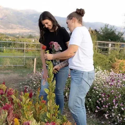 Two women picking flowers in a field with mountains in the background.