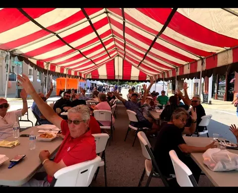 People sitting under a red and white striped tent, waving at the camera.