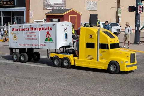 A small yellow toy-like semi truck with a trailer promoting hospitals drives down a street.