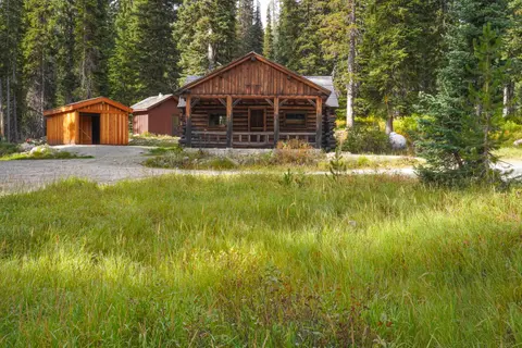 A rustic wooden cabin and outbuildings stand in a clearing surrounded by a dense forest.
