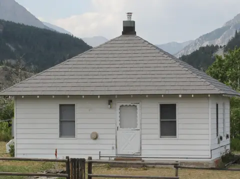 A small white building with a dark pyramid roof sits in a landscape with mountains.