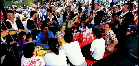 A mariachi band plays music outdoors near a crowd of people sitting at tables.