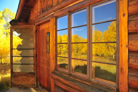 A rustic log cabin features windows reflecting autumn trees.
