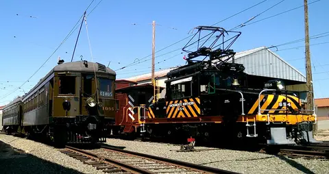 Two vintage electric trains sit on tracks outdoors under a clear blue sky.