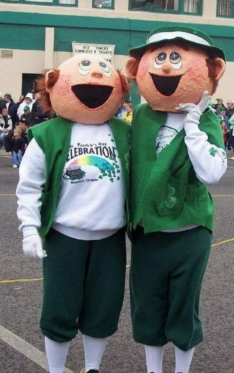 Two people wearing large costume headpieces and green outfits stand outdoors.