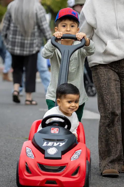 A young boy pushes a toy car with another child sitting inside it.