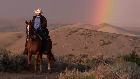 A person rides a horse across a grassy hillside with rolling hills and a rainbow in the background.