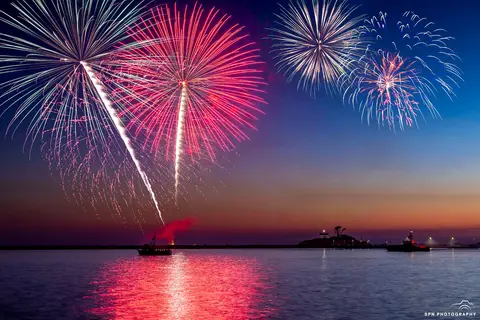 Colorful fireworks light up the night sky over a body of water with boats and a lighthouse in the distance.