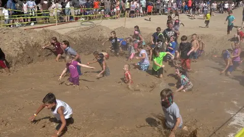 A large group of children play in a muddy pit while spectators watch from behind a yellow barrier.