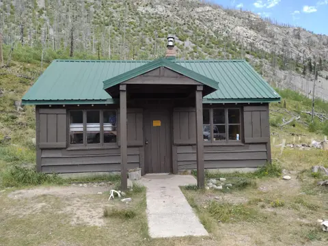 A small cabin with a green roof sits below a steep hillside.