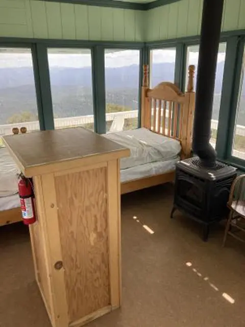 The interior of a cabin with a bed, wood stove, and large windows offering a scenic view.