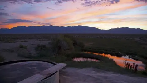A landscape photograph of a hot spring and mountain range during sunset.