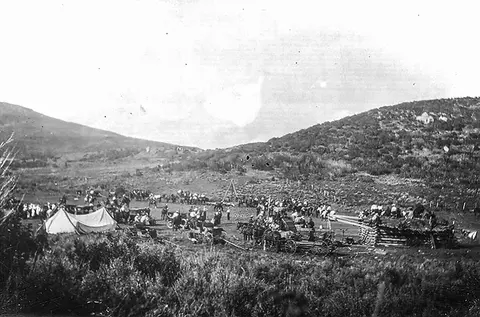 An old black and white photograph showing a gathering with tents and wagons in a valley between hills.