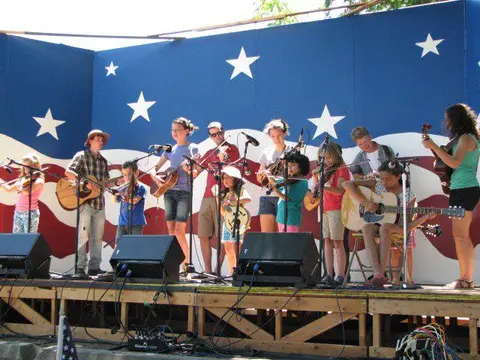 A group of young people playing musical instruments on an outdoor stage with an American flag backdrop.