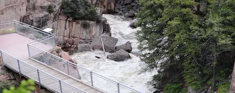 A wooden boardwalk and metal viewing platform overlooking a rushing river surrounded by rocks and trees.