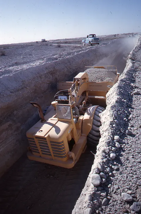 A heavy earthmoving vehicle operates in a long trench dug into a dry landscape.