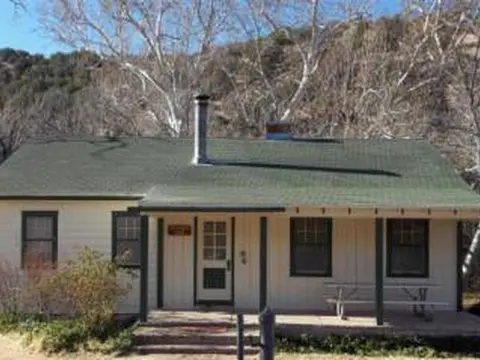 A single-story house with a green roof and a covered porch stands before a hillside and bare trees.