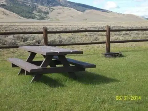 A wooden picnic table sits on a grassy lawn with a split-rail fence and mountains in the background.