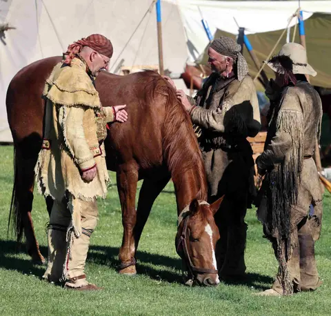Three people in historical frontier clothing stand around a grazing horse outdoors.