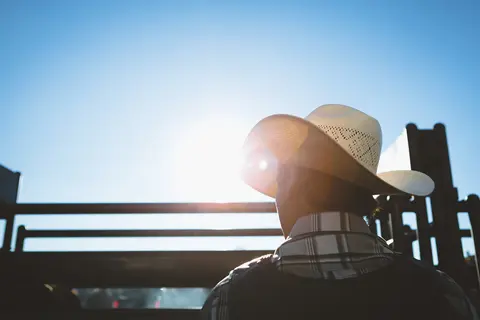 A person wearing a cowboy hat stands near a wooden fence against a bright sky.