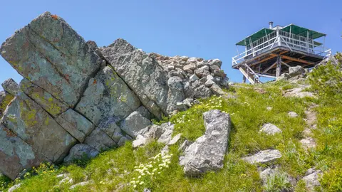 A fire lookout tower sits atop a rocky, grassy mountain peak under a clear blue sky.