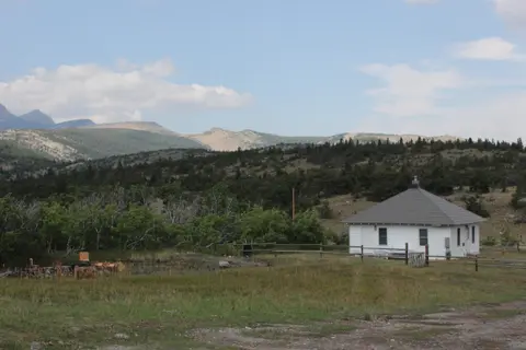 A small white building sits in a grassy field with mountains in the background.