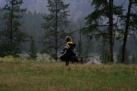 A person running through a grassy field with trees and mountains in the background.
