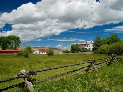 A rustic wooden fence runs across a grassy field with rural buildings and a cloudy sky in the background.
