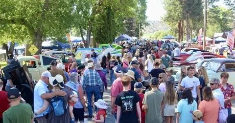A large crowd of people walking along a street lined with vintage cars and trees.
