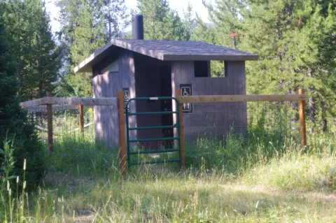 An outdoor toilet building is surrounded by a wooden fence in a wooded area.