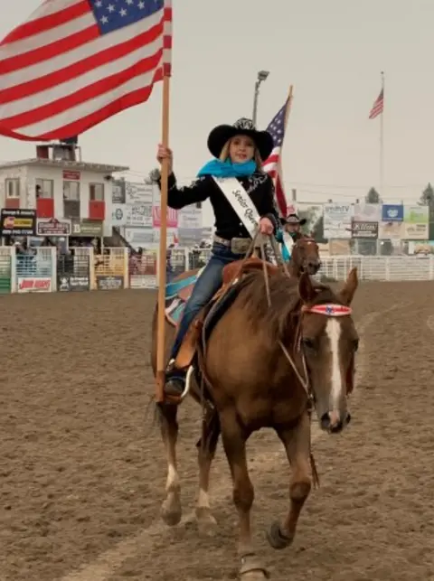 A young woman on a horse holds an American flag in an arena.