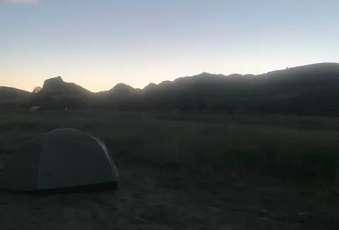 A tent sits in a field beneath distant silhouetted hills at sunrise.