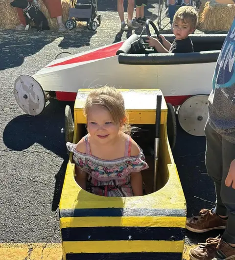 A young girl sits in a small yellow wooden toy car outdoors.
