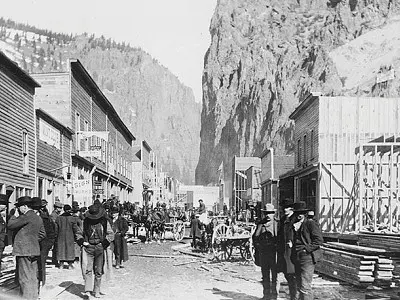 Historic photo showing a busy dirt street in a frontier town with wooden buildings and mountains in the background.