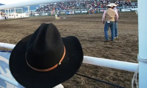 A black cowboy hat rests on a fence overlooking a rodeo arena with spectators in the background.