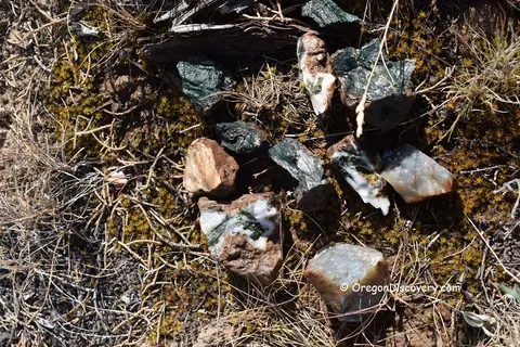 A collection of raw stones and minerals resting on dirt and dry grass.