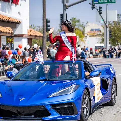 A woman in a red dress with a sash and tiara waves from a blue convertible in a parade.