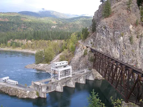 A dam and a railway bridge span a river next to a rocky cliff and forested hills.