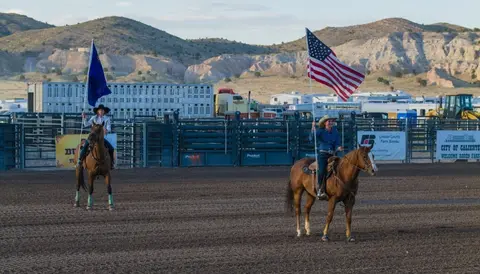 Two women on horseback carry flags inside a rodeo arena.