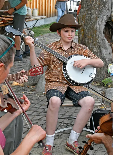 A young boy plays a banjo while sitting outdoors with other musicians.