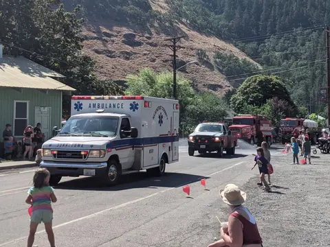 An ambulance and fire trucks drive down a street with people watching.