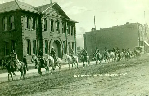 A historic black and white photo shows people on horseback riding past a brick building.