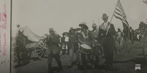 An old photograph showing a group of people marching in a procession with an American flag.