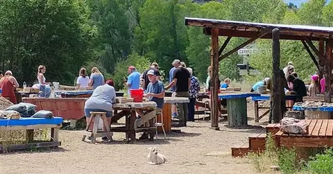 People at outdoor tables under a wooden shelter and a small dog sitting on the ground in the foreground.