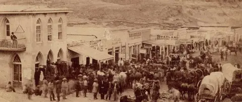 A historic photograph showing a busy street in an old town with buildings and horse-drawn wagons.