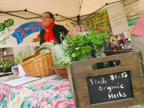 A vendor stands behind a market stall displaying fresh herbs and vegetables.