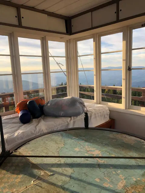 An interior view of a fire lookout tower with large windows showing a mountain landscape.